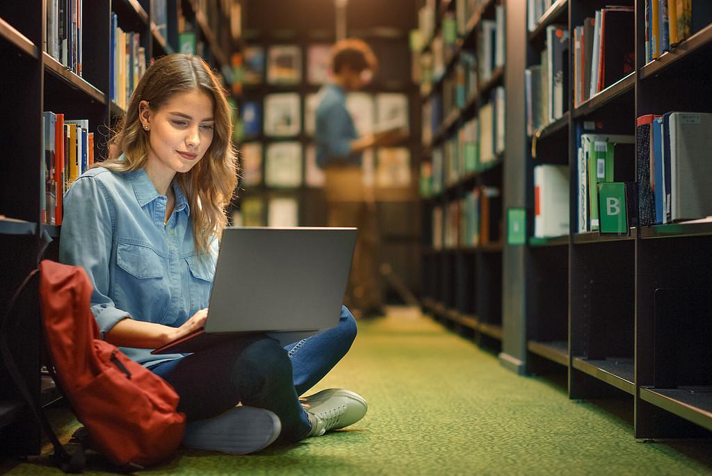 Eine Promovierende sitzt zwischen zwei Bücherregalen auf dem Boden der Bibliothek. In ihrem Schoß liegt ihr aufgeklappter Computer, in dem sie zu den Richtlinien der guten wissenschaftlichen Praxis recherchiert.