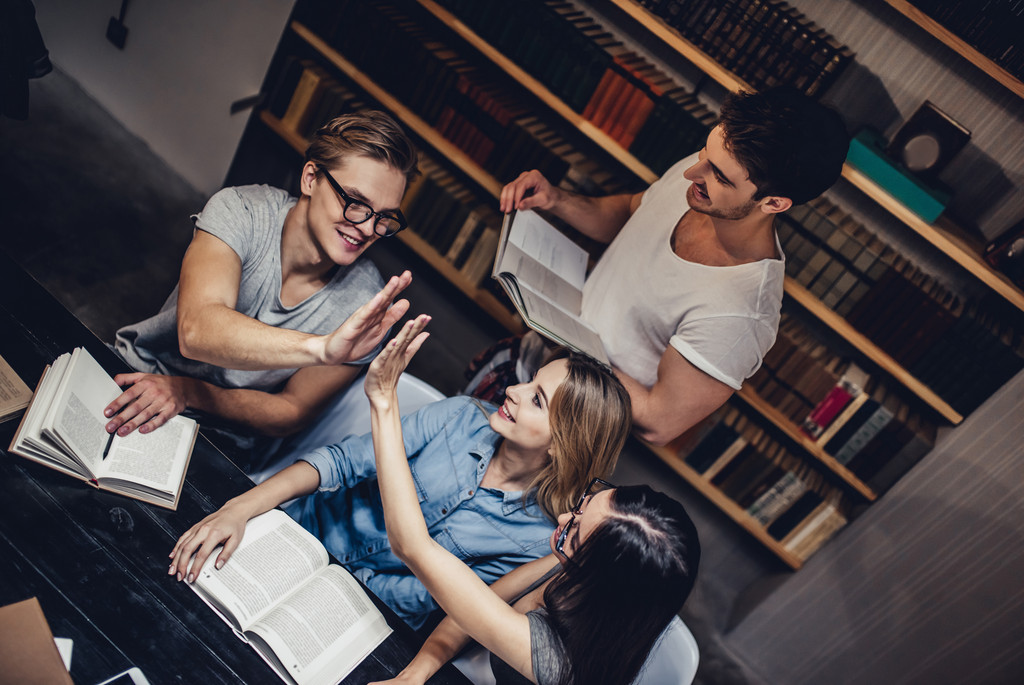 Drei Studierende sitzen über Büchern an einem Tisch in einer Bibliothek, zwei klatschen sich gegenseitig zu. Hinter ihnen steht ein weiterer Student mit einem aufgeschlagenen Buch in der Hand.